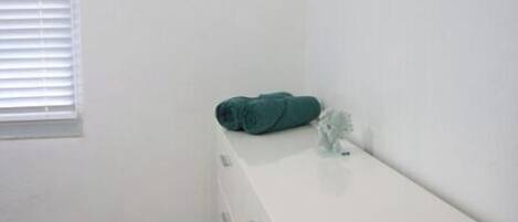 Une petite pièce propre, avec une commode blanche, une fenêtre équipée de stores et un radiateur blanc.