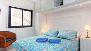 Ein Schlafzimmer mit einem Bett, zwei Nachttischlampen, einem Korbstuhl und einem Fenster mit Blick auf kahle Bäume.