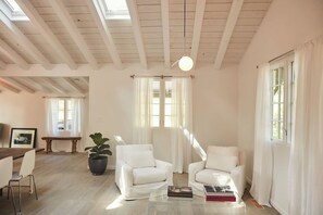 Une pièce spacieuse avec un plafond haut, des murs blancs et une grande fenêtre ornée de rideaux blancs. On y trouve un banc en bois, une plante en pot et deux fauteuils blancs disposés autour d’une table basse.