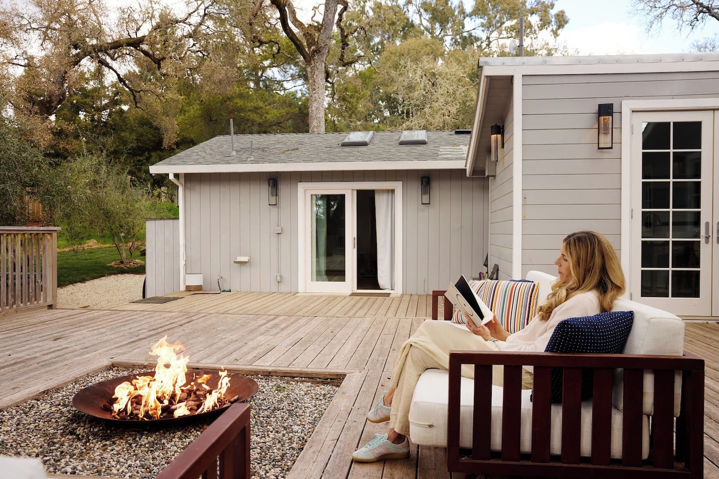 A person sitting on a deck chair reading a book, with a fire pit in the foreground and a building with a sliding door in the background.