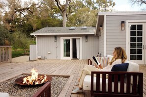 A person sitting on a deck chair reading a book, with a fire pit in the foreground and a building with a sliding door in the background.