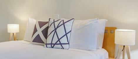A neatly made bed with white linens and two decorative pillows, flanked by bedside lamps on wooden nightstands.