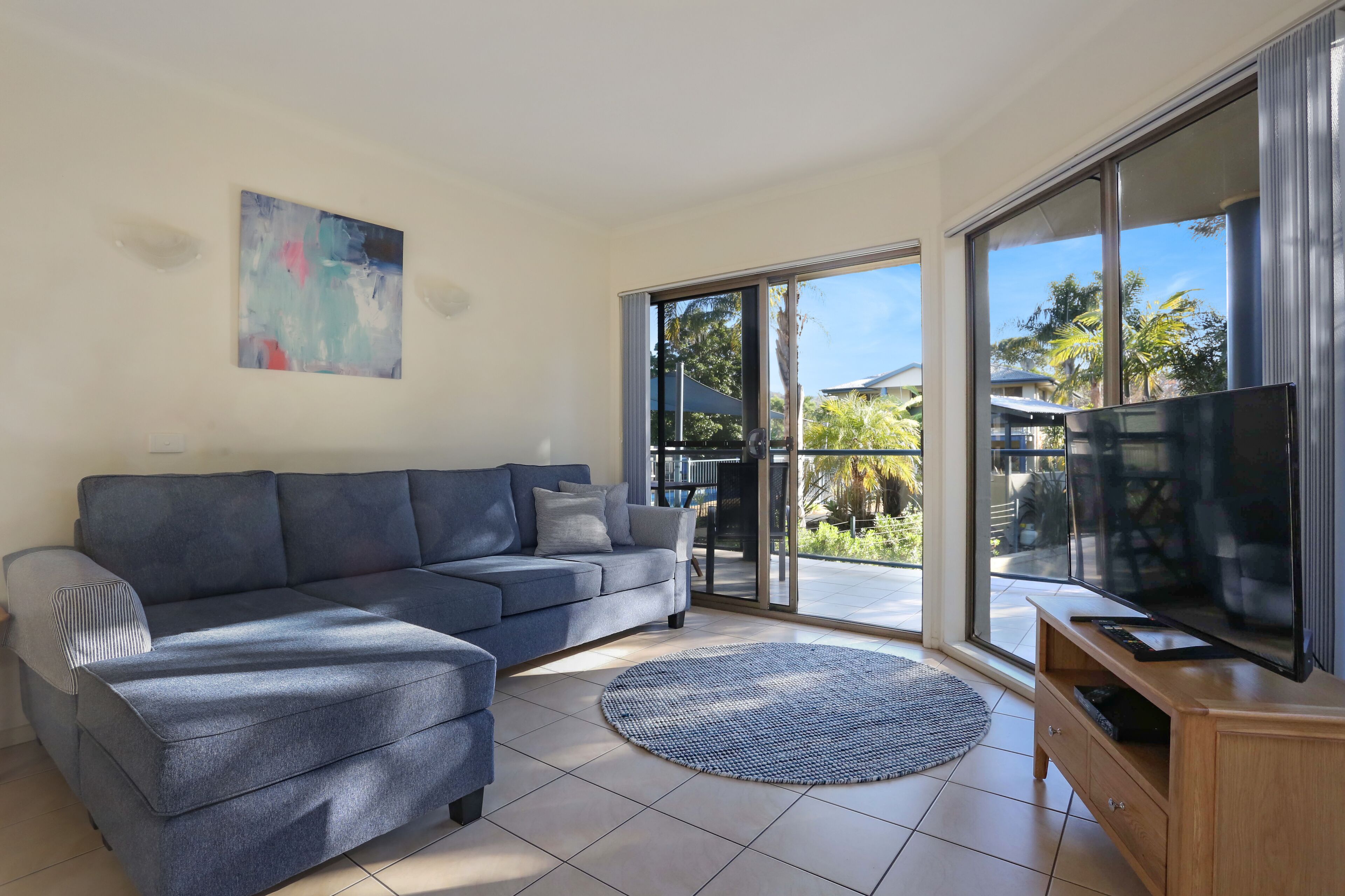 A modern living room with a sectional sofa, a flat-screen TV, and a large sliding glass door leading to a balcony with outdoor furniture.