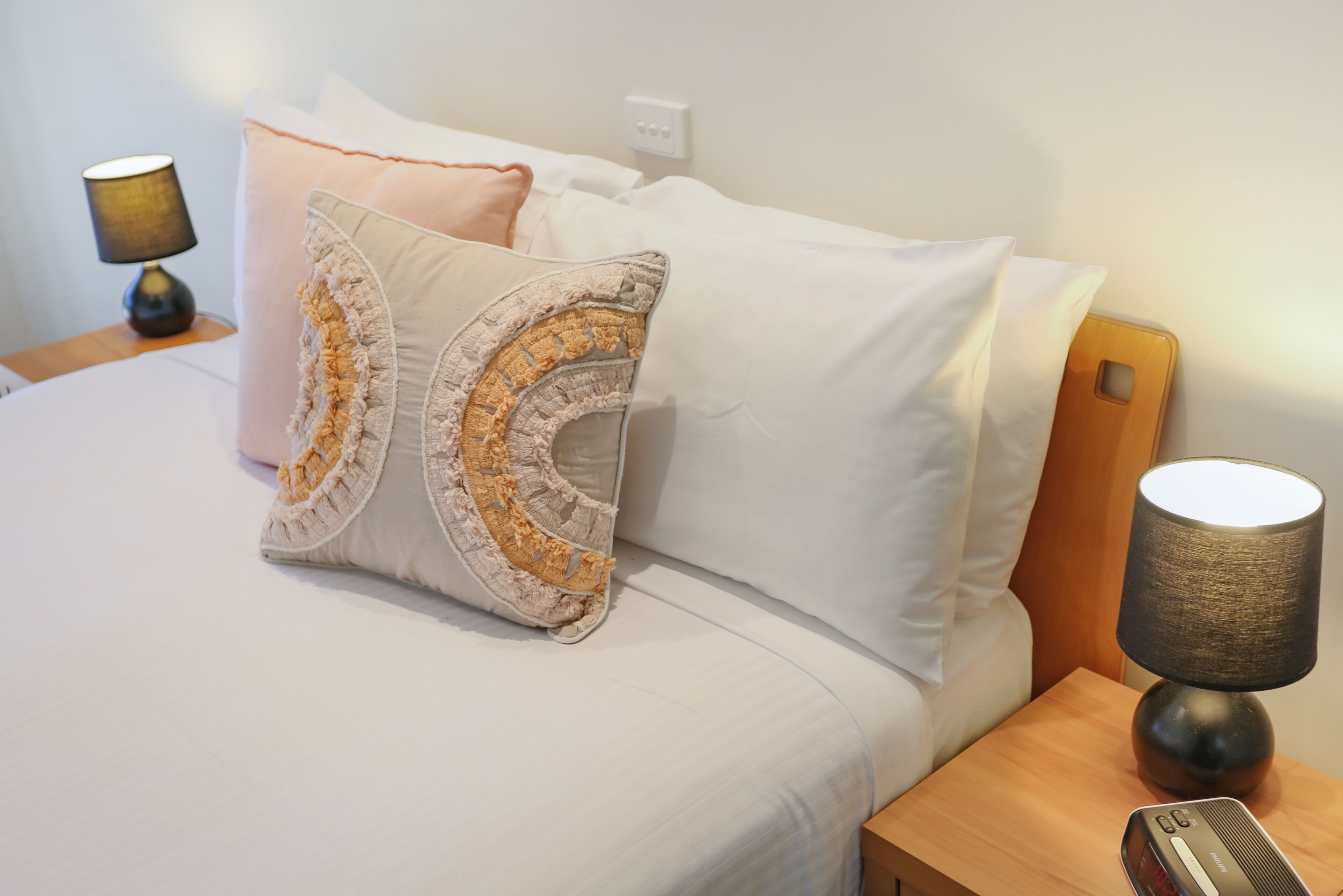 En pænt redt seng med hvide sengetøj og dekorative puder, et sengebord med en lampe og et digitalt ur.