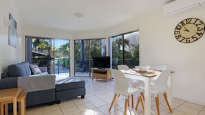A modern living room with a sofa, TV, dining table, and chairs. - Sorrento Apartments Merimbula (Merimbula)