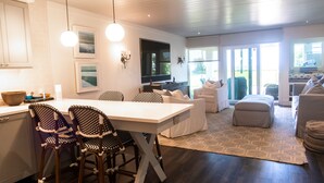 Una cocina moderna con comedor, una sala de estar con sofá y reposapiés, y un amplio ventanal con vistas al exterior.