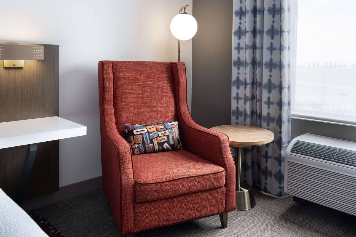 A red armchair with a patterned cushion, a small round table, a white bedside table, a wall-mounted light, and a window with curtains.
