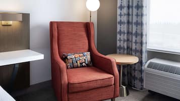 Un fauteuil rouge avec un coussin à motifs, une petite table ronde, une table de chevet blanche, une lampe fixée au mur et une fenêtre avec des rideaux.