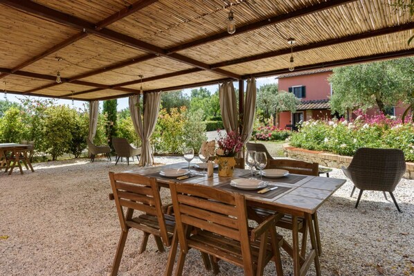 An outdoor covered dining area with tables set for two, surrounded by gravel, with a view of a garden and a building in the background.