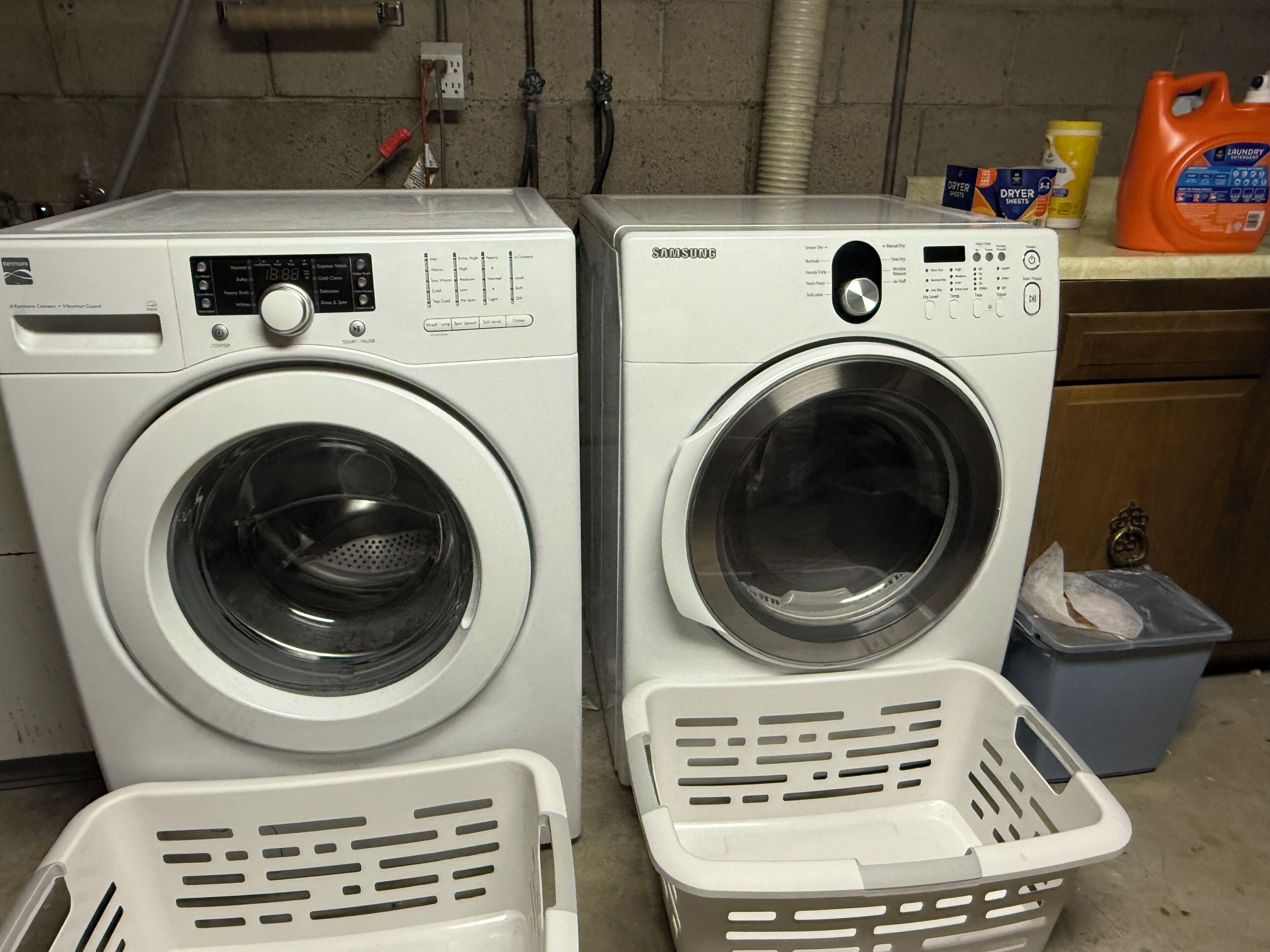 Two front-loading washing machines in a laundry area with a blue bin and a yellow container in the background.