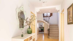 En gang med et hvidt konsolbord, en grøn vase med tørrede blomster, et spejl med blomsterprydet ramme, og en trappe, der fører til et øvre plan.
