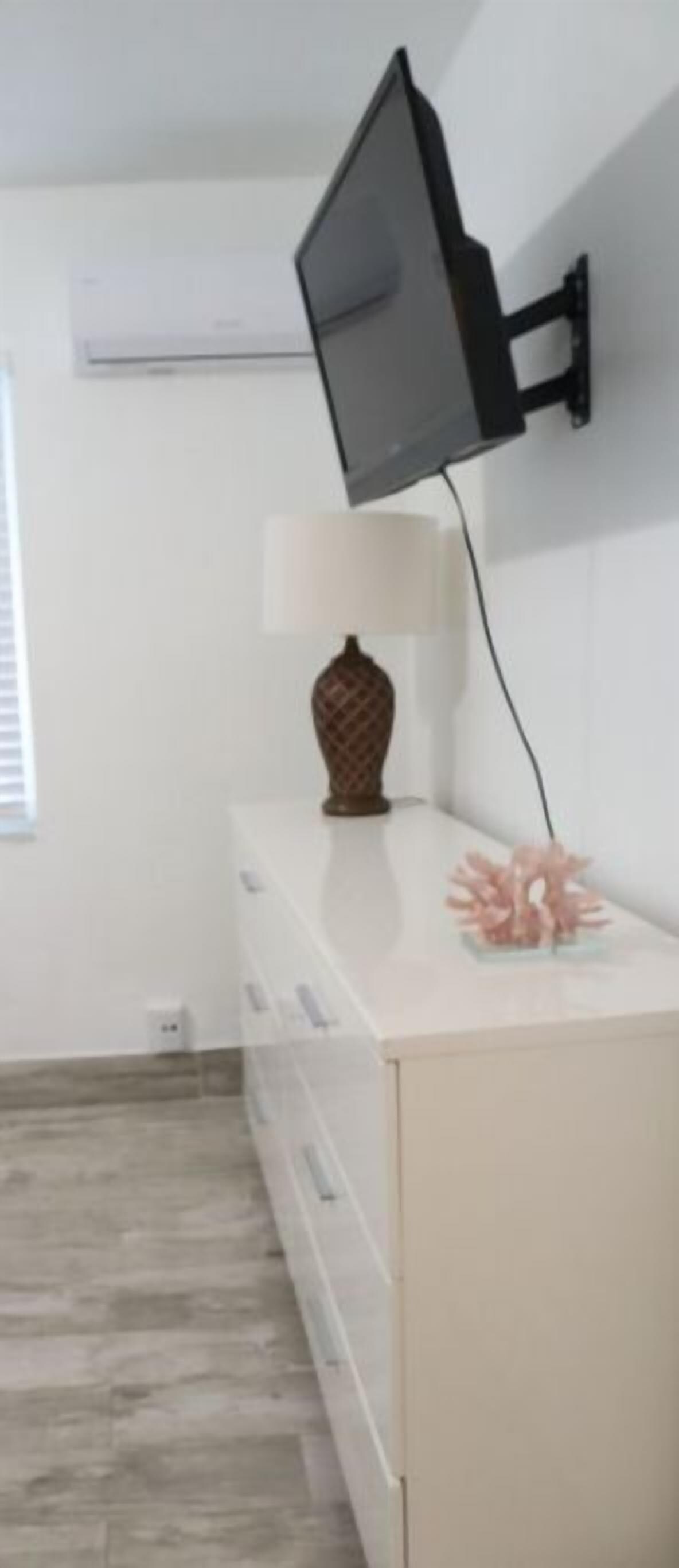 A room with a mounted TV, a white dresser, a vase, and a pink decorative item.