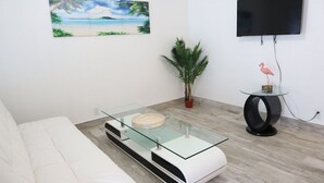 A modern living room with a glass coffee table, a white sofa, a potted plant, a small table with a flamingo figurine, and a flat-screen TV mounted on the wall. - WoW!! Stunning 1 BR // Prime Singer Island (West Palm Beach)