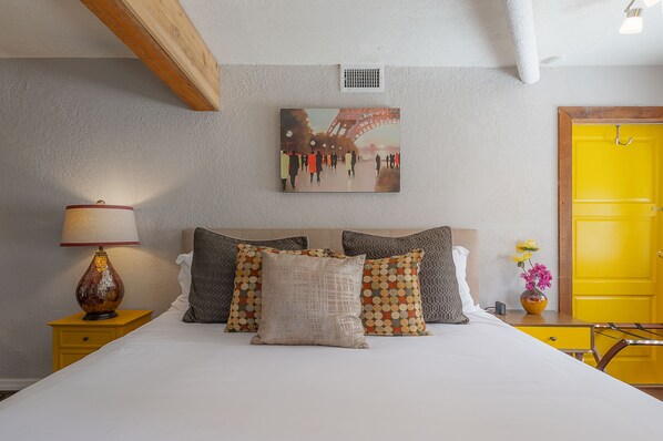Une chambre d’hôtel avec un lit, des oreillers, une table de chevet, une lampe, un tableau, un vase avec des fleurs et une porte jaune.