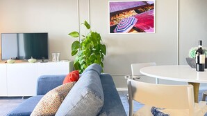 Ett modernt vardagsrum med en blå soffa, ett vitt matbord, en krukväxt och en tv-apparat med platt skärm.
