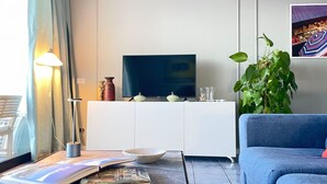 Ett modernt vardagsrum med en stor tv-skärm, ett vitt skåp, en blå soffa, ett soffbord i glas och en krukväxt.