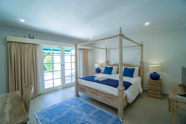Una cama con dosel de madera, con colcha y almohadas azules y blancas. En el suelo, hay una alfombra azul, una mesita de noche de madera con una lámpara, y una silla.