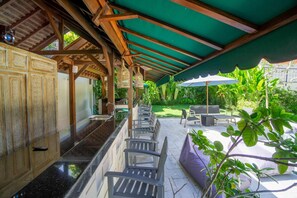 A covered outdoor seating area with chairs and tables, a bar counter, and a view of a lush garden.