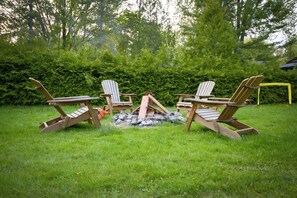 En uppsättning Adirondack-stolar är placerade runt en eldstad på en gräsig utomhusplats, med tät vegetation i bakgrunden.