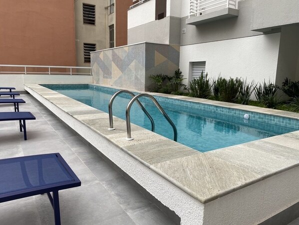Una piscina con plataforma de baldosas, barandillas de metal y algunas sillas de descanso.