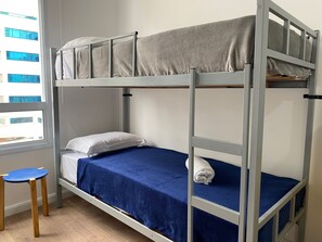 Una litera con colchón azul y manta gris. Junto a la cama hay un pequeño taburete de madera.