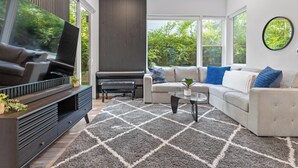 Una sala de estar moderna con un televisor grande, sofá y mesa de centro, con un rug geométrico y ventanas amplias con vistas a zonas verdes.