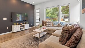 Una sala de estar moderna con un televisor de pantalla plana grande, un mueble blanco para equipo multimedia, un sofá color beige, cojines estampados y un ventanal con vistas a la vegetación.