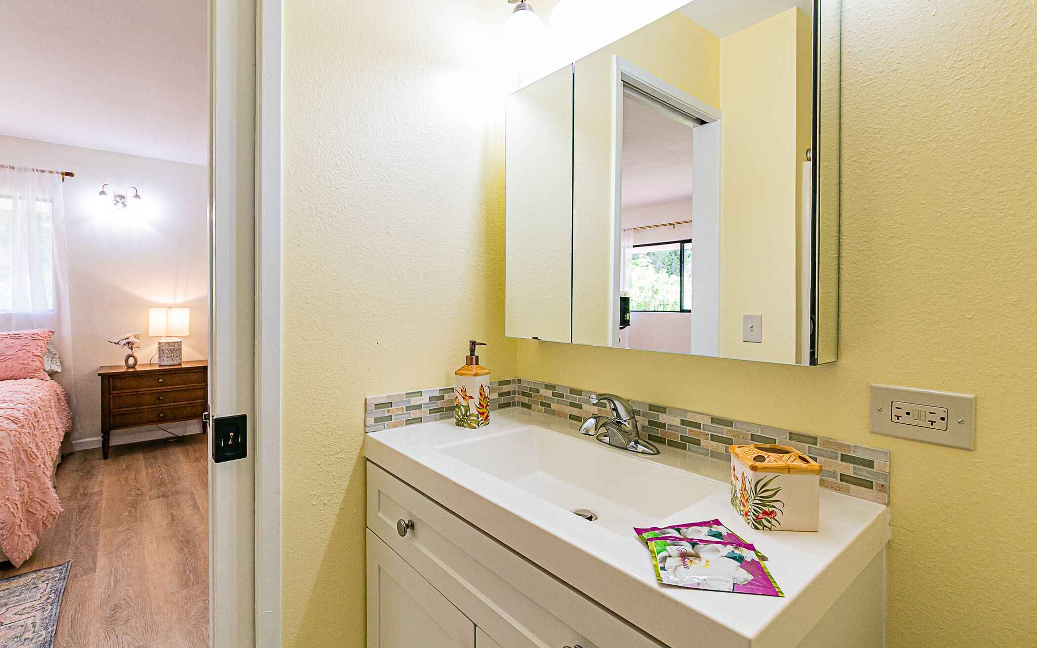 A bathroom with a white sink, a mirror, and a tiled backsplash. There is a small cabinet below the sink and a door leading to another room.