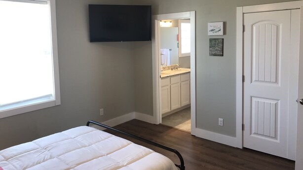 Une chambre avec un lit, un ventilateur de plafond, un téléviseur fixé au mur, une salle de bain avec un lavabo et un miroir, et une porte ornée d’une plaque décorative.