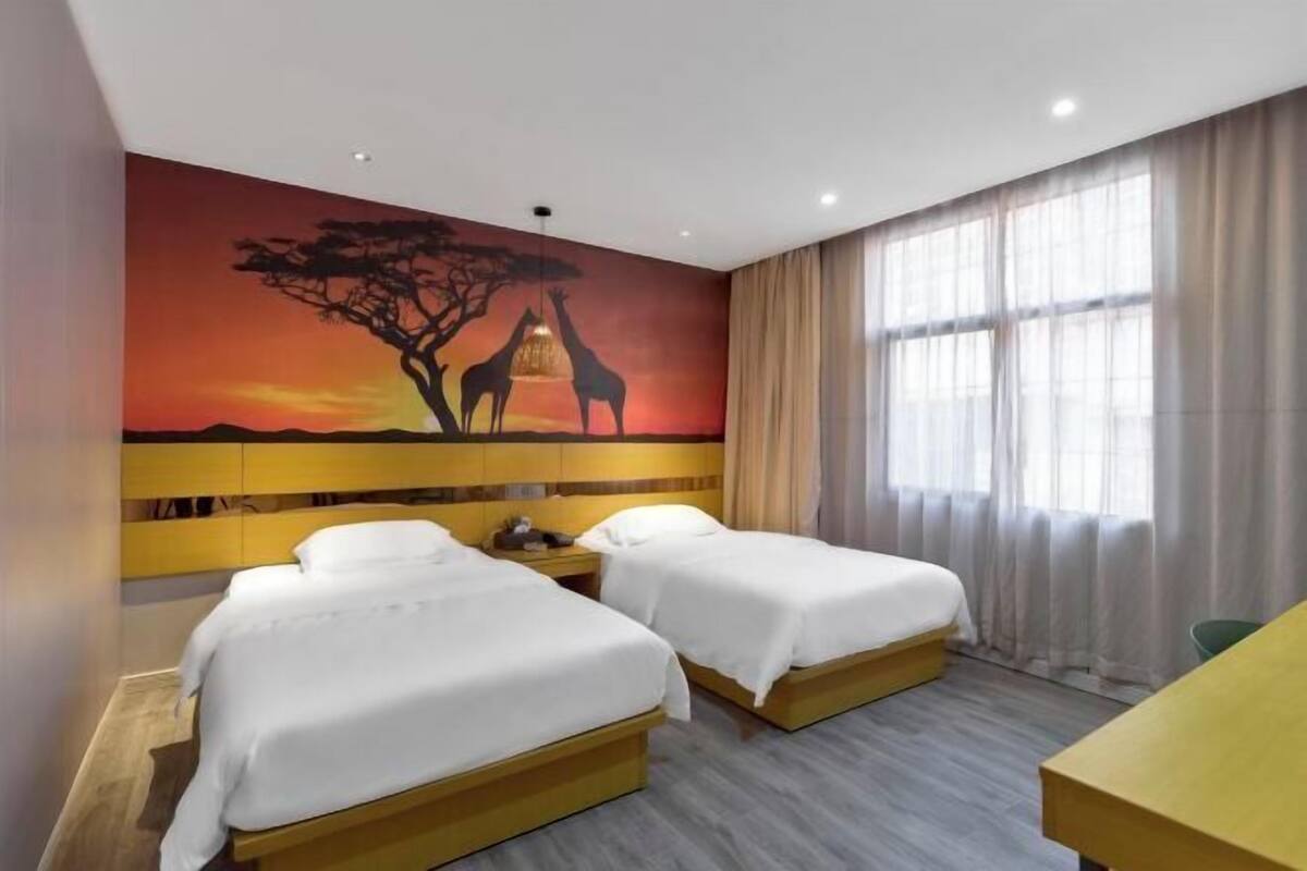 A hotel room with two beds, a painting of giraffes, a desk, and a window with curtains.