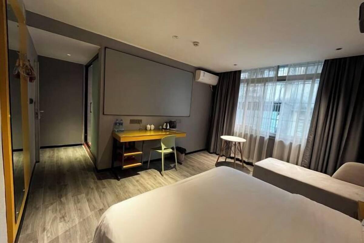 A hotel room with a bed, a desk, a chair, a small table, and a large window with curtains.