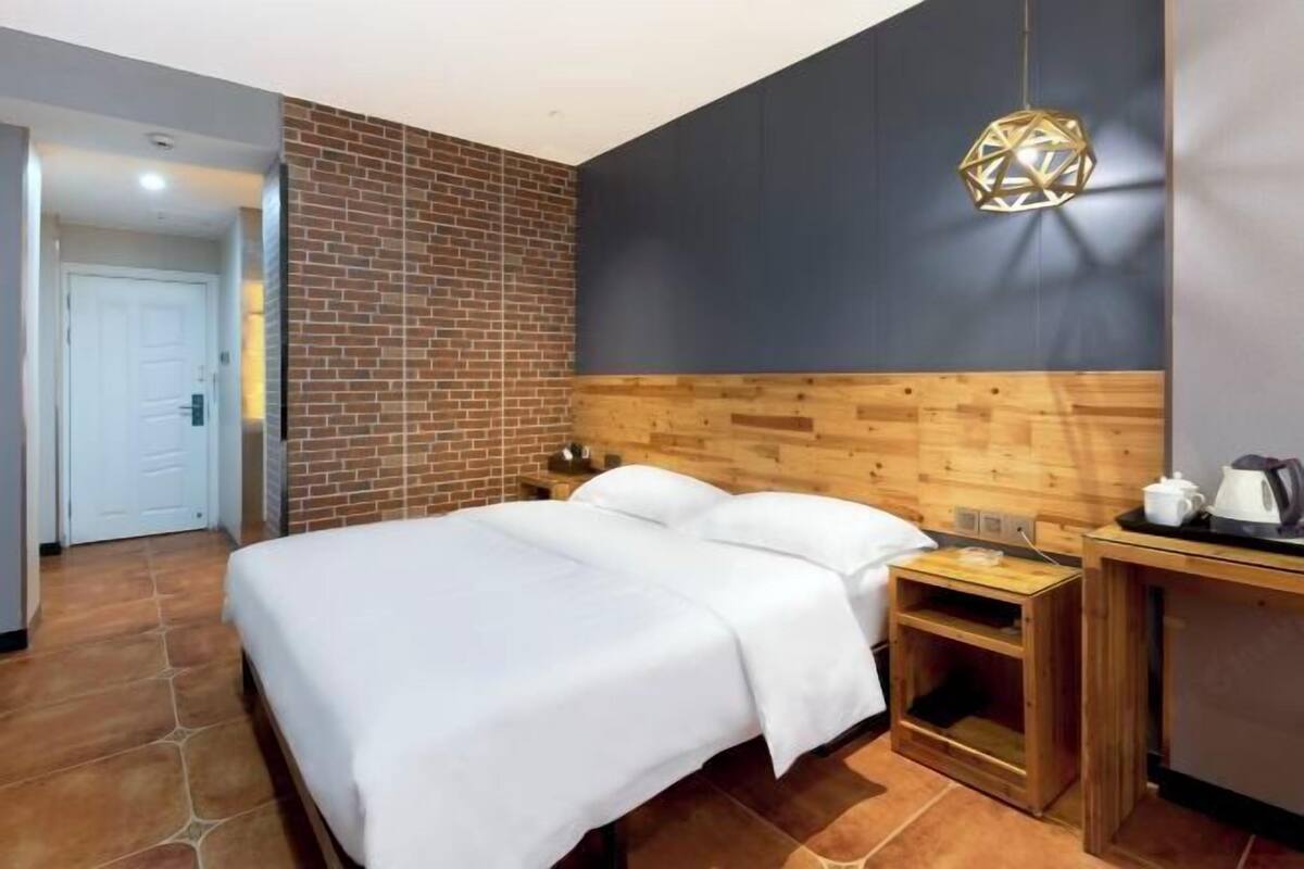 A hotel room with a brick accent wall, a wooden headboard, a bed with white linens, a small wooden nightstand, and a hanging light fixture.