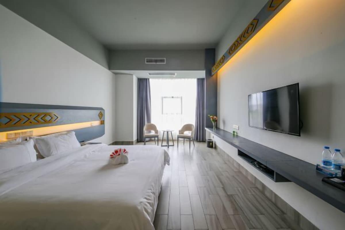 A hotel room with a large bed, a TV mounted on the wall, a small seating area with two chairs, and a window with curtains.