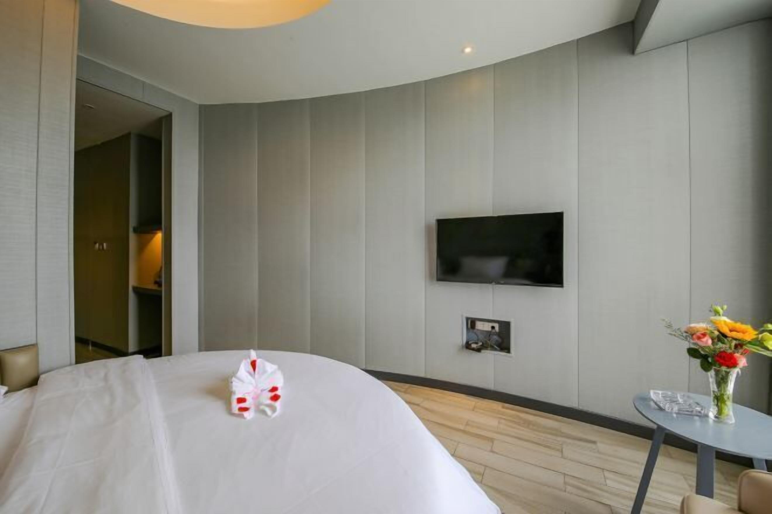 Moderni hotellihuone, jossa on suuri sänky, seinälle asennettu taulutelevisio, pieni pöytä maljakko täynnä kukkia ja ovi, joka johtaa toiseen huoneeseen.