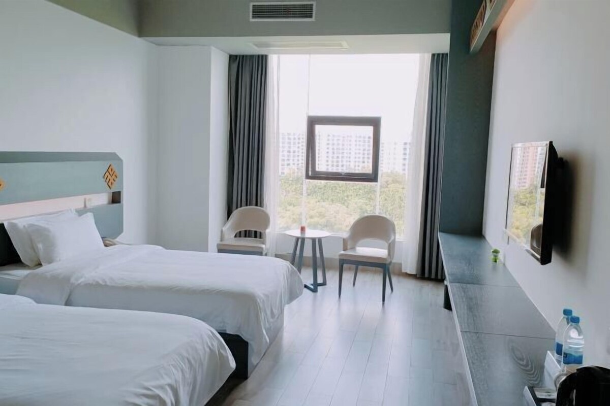A hotel room with two beds, a small table, two chairs, a TV, and a window with a view of buildings.