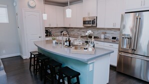Cocina moderna con una isla central, taburetes de bar, electrodomésticos de acero inoxidable y un reloj en la pared.
