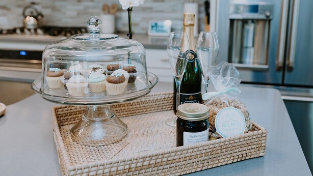 Una encimera de cocina con un soporte de vidrio para pasteles con cupcakes, una botella de champán y un frasco de mermelada.