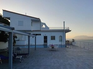 Ein modernes, weißes Gebäude mit überdachter Terrasse, Sitzgelegenheiten im Freien und einer Glaskonstruktion.