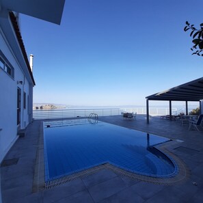 Ein Schwimmbad mit Meerblick, eine überdachte Terrasse mit Sitzgelegenheiten und ein Gebäude mit weißer Fassade.