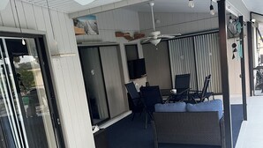Un espace extérieur aménagé en patio, doté de ventilateurs de plafond, d’un coin salon avec un canapé et des fauteuils, et d’un téléviseur.