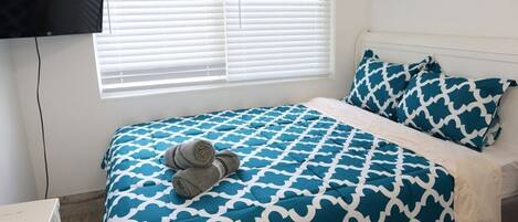 A bedroom with a bed, a TV mounted on the wall, and a window with blinds.