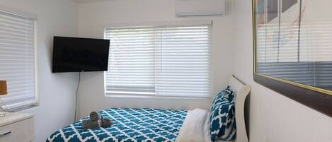 A bedroom with a bed, a TV mounted on the wall, and a window with blinds.