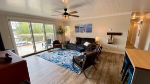 Un salon spacieux doté d’une grande fenêtre, d’un ventilateur de plafond, d’un canapé en cuir noir, d’une table basse et d’un tapis bleu.