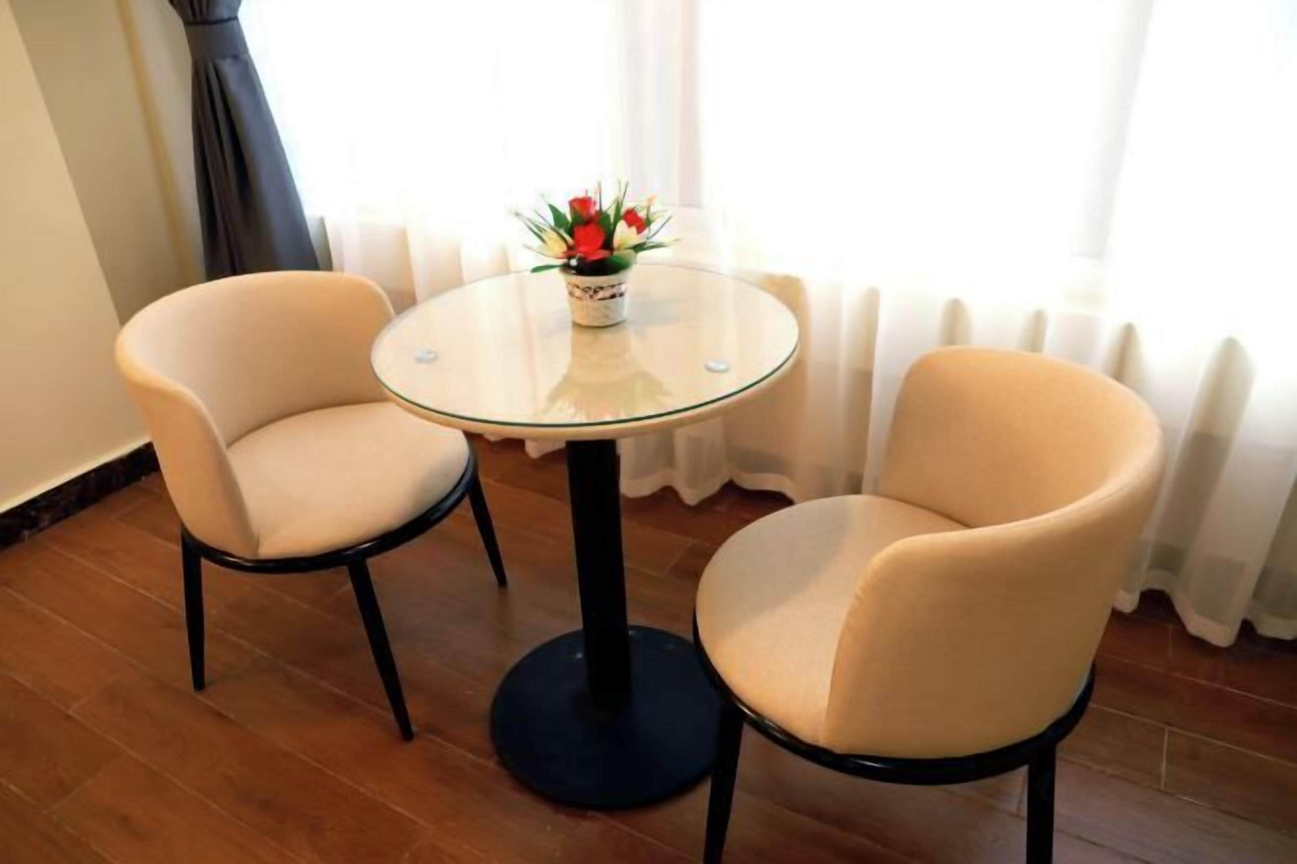 A room with two beige chairs around a small glass table, a potted plant on the table, and a window with curtains.