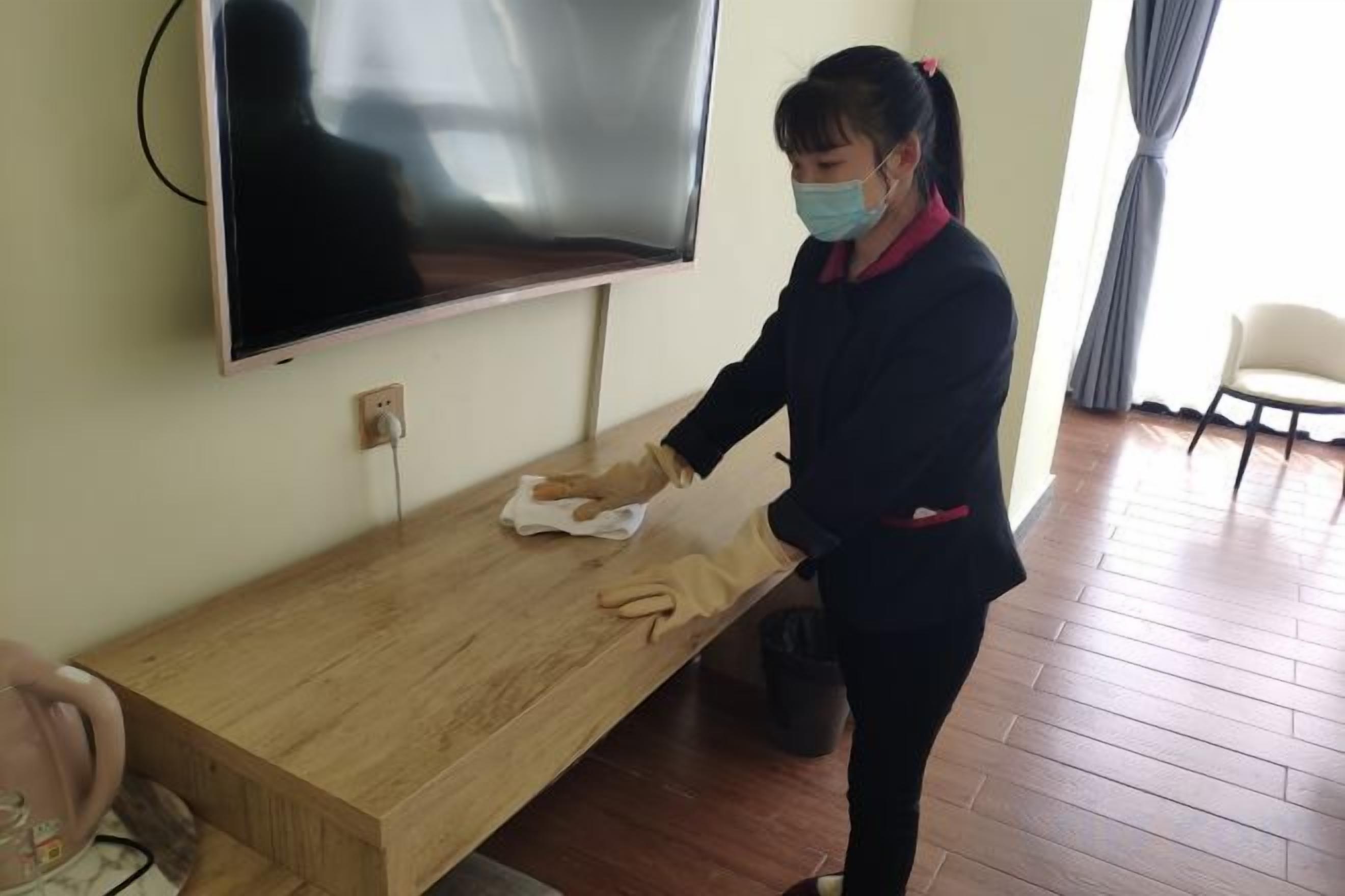 Una persona limpiando una mesa de madera en una habitación con un televisor montado en la pared y una silla al fondo.