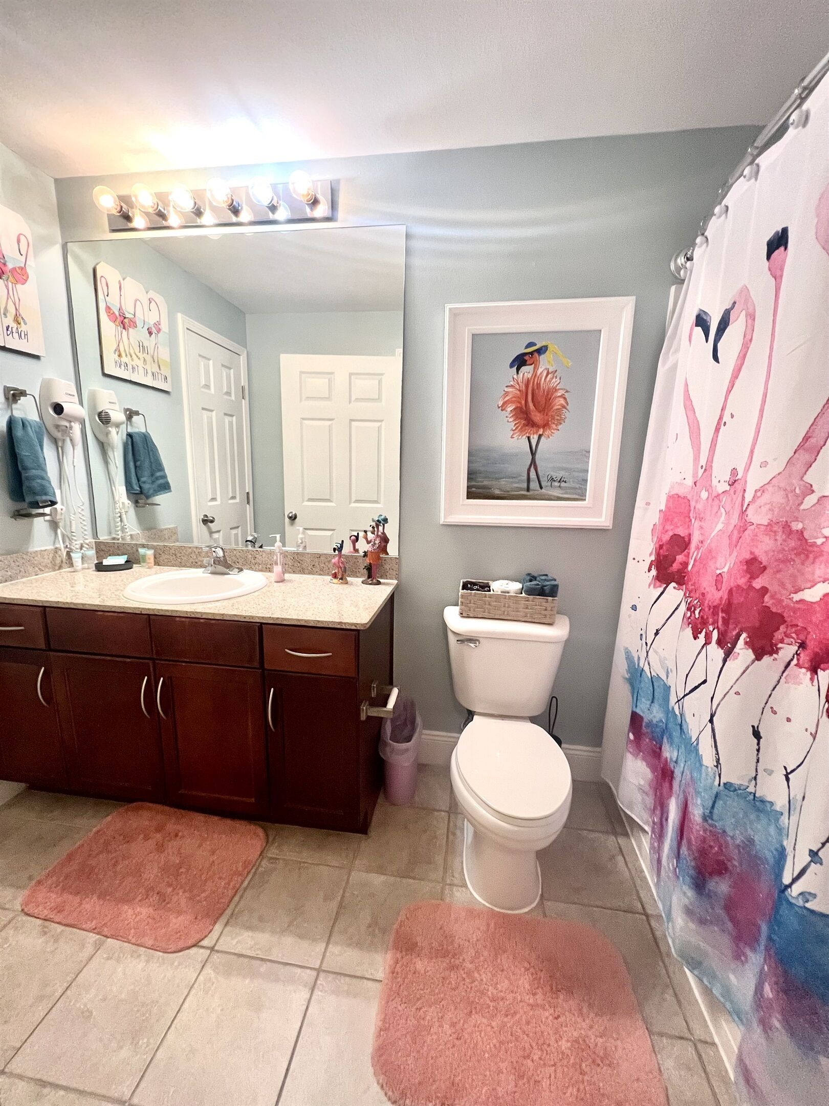 Une salle de bain avec un meuble-lavabo, des toilettes, un rideau de douche orné de motifs floraux et un tableau encadré représentant un flamant rose accroché au mur.