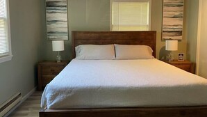 Een slaapkamer met een houten hoofdbord, een groot bed met wit beddengoed, twee nachtkastjes met lampen en twee aan de muur hangende schilderijen.