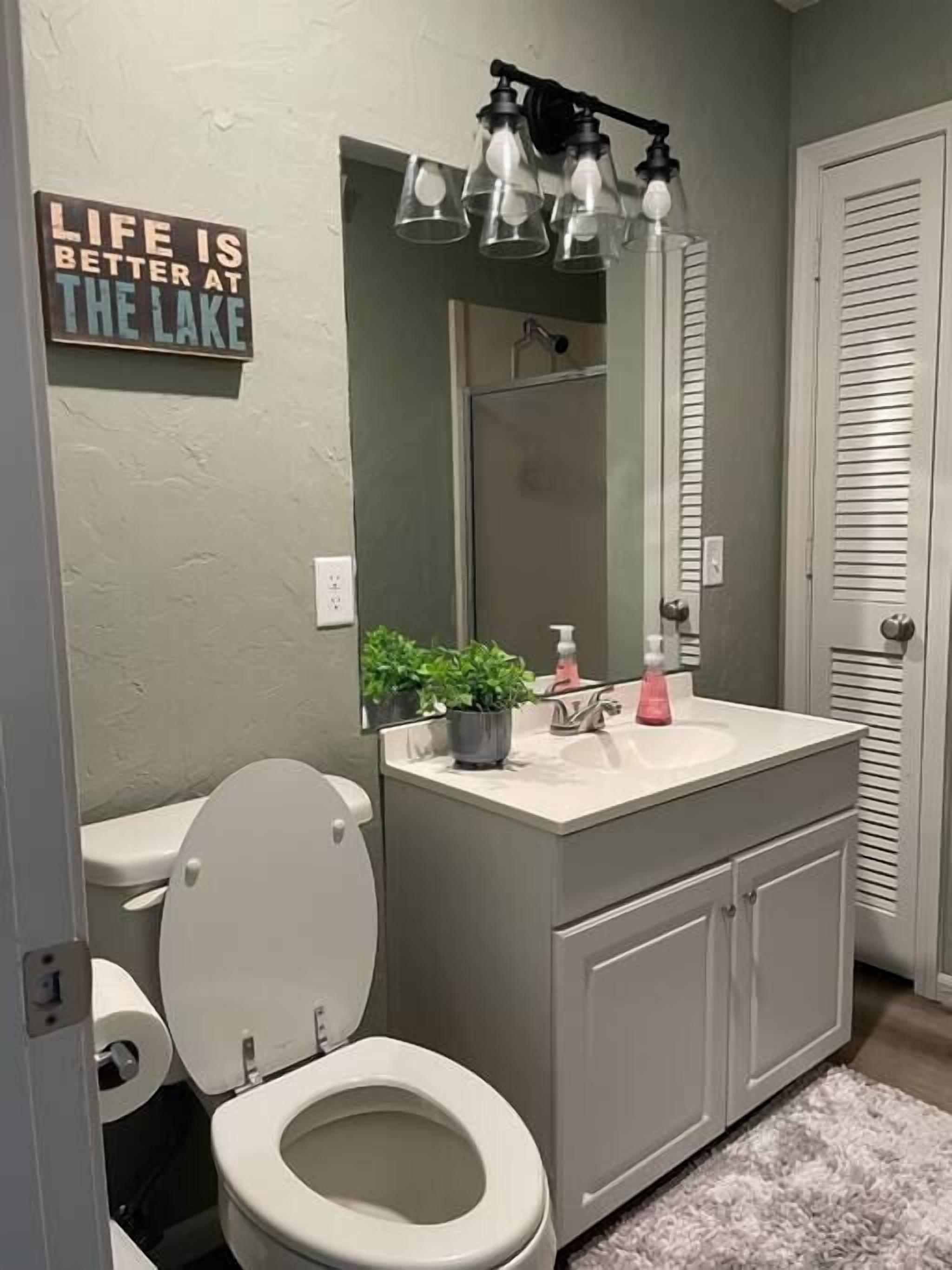 A bathroom with a toilet, sink, and mirror. There is a sign on the wall that reads "Life is better at the lake."