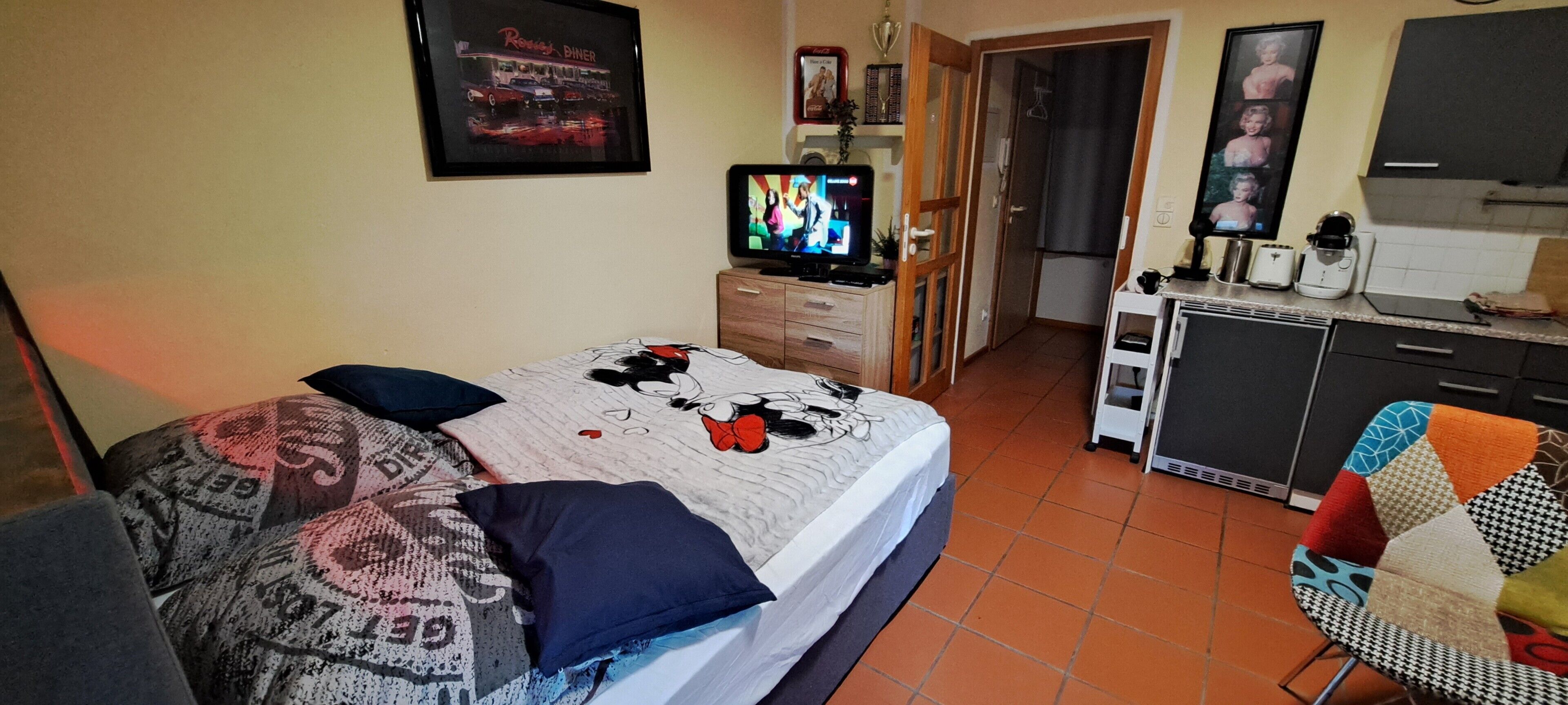 A bedroom with a bed, a television, a dresser, and a kitchen area with a refrigerator and microwave.