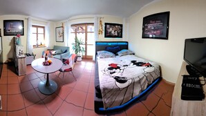 Un espace de vie compact comprenant un coin cuisine, un coin repas, un lit avec une couverture aux motifs de Mickey Mouse, une télévision et une plante.